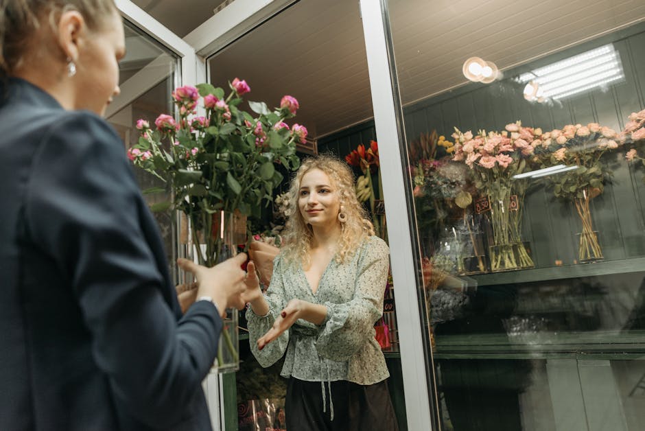 A florist, with blonde, curly hair and wearing a patterned blouse, is in a flower shop behind glass doors, receiving a bouquet of fresh pink roses with green stems and leaves wrapped in brown paper fr