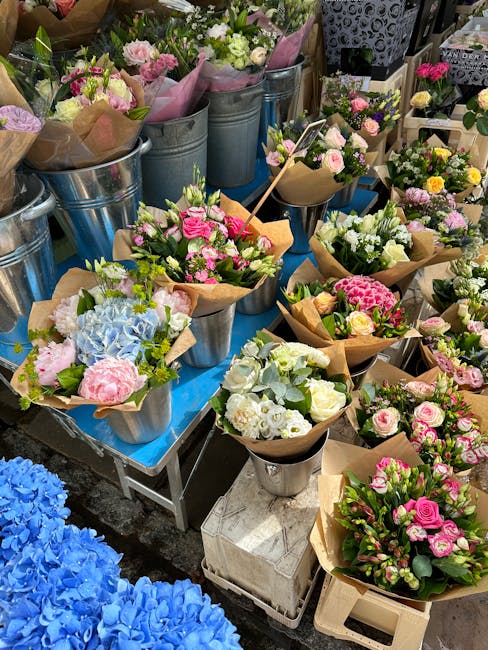 An assortment of fresh flower bouquets displayed on a table outdoors, featuring a variety of pastel pinks, whites, and blues. The arrangements include roses, hydrangeas, and other seasonal blooms, wra