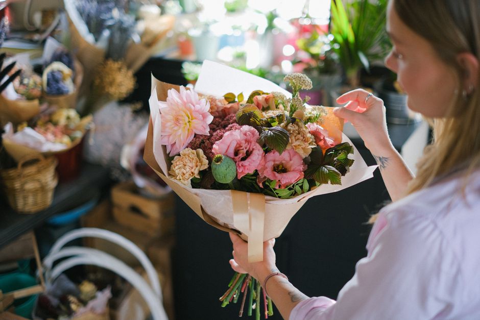 A woman with light brown hair, wearing a pale pink shirt, is holding a bouquet of fresh flowers in a florist shop. The bouquet features pink and cream dahlias, pink lisianthus, and assorted green foli