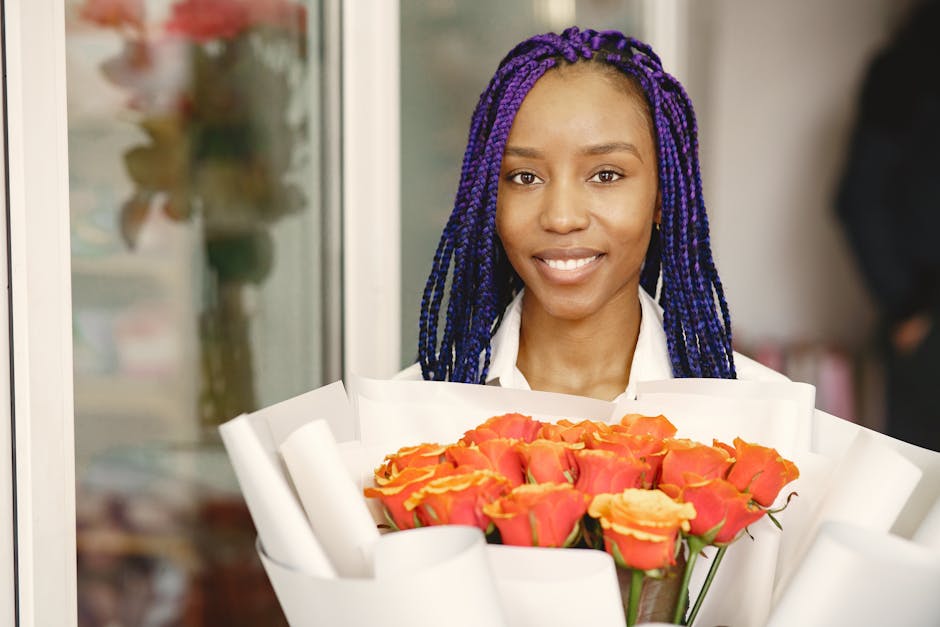 A young woman with dark skin and long, vibrant purple braided hair is standing outdoors, holding a large bouquet of fresh roses arranged in a spiral pattern. The roses are primarily coral and peach in