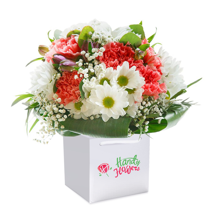 Presented in a snow-white, geometric gift bag that sits squarely against a clear white backdrop, this vibrant bouquet is a celebration of colour and careful design, just as you might find in South Woodford's welcoming local flower shops. The heart of the arrangement is formed by lush, ruffled-petal carnations in shades of coral and red, spilling warmth and a textured abundance, as if freshly gathered from garden beds near Wanstead Flats. Sprays of crisp white daisies with lime-green centers peek playfully throughout, providing a fresh, uplifting contrast akin to daisy clusters in Elmhurst Gardens. The bouquet's surface glimmers with soft, cloudlike tufts of baby's breath, creating subtle dimension and a hint of gentle movement. Along the left, several petite unopened buds with purple blushes add a point of visual interest and the promise of blooms yet to come. All of these are cocooned within radiant, dark green foliage-broad, glossy leaves that curve around the composition, offering support and drawing the eye inward. The minimalist elegance of the white bag, topped with coordinating handles, makes the arrangement feel equally perfect for stylish home décor or as a memorable gift, ready for same-day delivery across South Woodford and neighbouring areas. This detailed portrayal captures the freshness and premium quality that South Woodford Florist delivers, reflecting both their artistry and deep community ties.