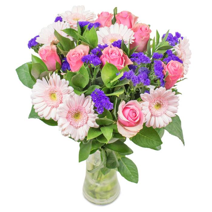 A vibrantly styled floral bouquet sits in a sleek glass vase, its reflection set against a crisp, white background-the kind often seen in homes around South Woodford. At its core, 7-9 pink roses, their petals tightly furled or just beginning to unfurl in perfect spiral patterns, rise gracefully between 5-7 pale, blush-pink gerbera daisies. Each gerbera displays wide, radiating petals with gentle, almost white centres, echoing the freshness of Woodford's village green in early summer. Interspersed, clusters of vivid purple statice provide bold textural dimension and lively colour contrast, reminiscent of a stroll through the Sunday market flower stalls. The lush, deep-green foliage forms a dense, abundant base, infusing the bouquet with the look and scent of the forest edges bordering the Roding Valley. The transparent vase highlights sturdy stems and clear water, finishing the scene with a pure, natural aesthetic. This bouquet marks joyful occasions-birthdays, anniversaries, or as a thoughtful gift-celebrating affection, local charm, and the artful curation that South Woodford residents have come to expect from their favourite floral studio.